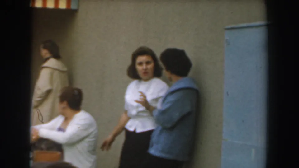 1961 Los Angeles home-movie frame showing two standing women talking beside a table with seated adults.