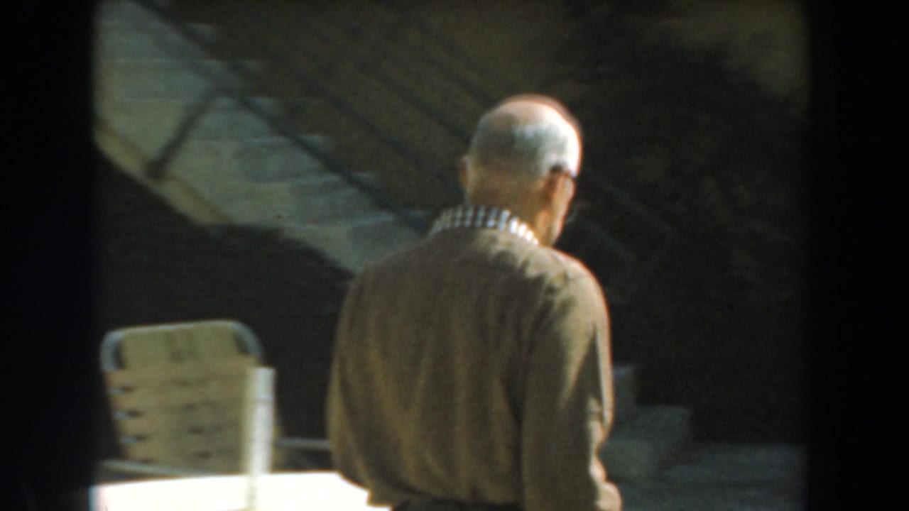 Adult man with glasses seen from behind near a patio chair and shaded exterior wall.
