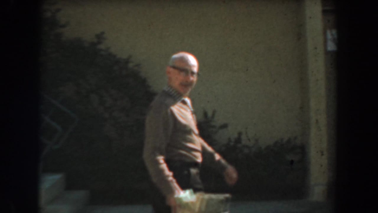 Adult man with glasses walking past bushes while holding a small box or container.