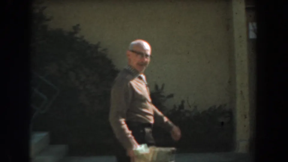 In 1961 Los Angeles, a man in glasses and a brown shirt walks through a residential yard carrying a small box or container.