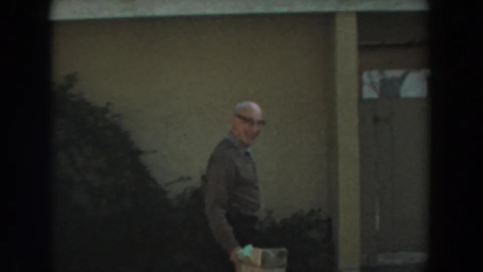 The man with glasses continues walking along the residential exterior, with bushes, a doorway, and textured wall visible behind him.