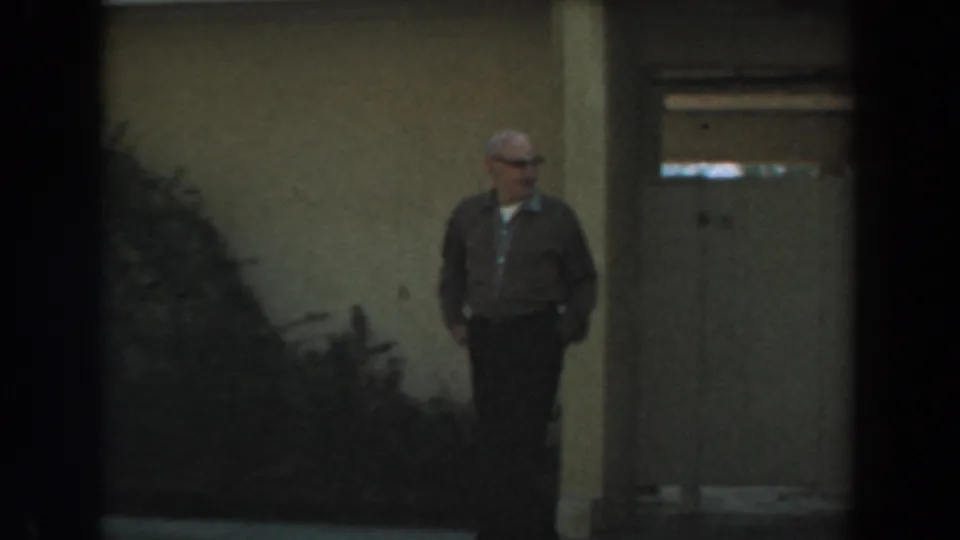 A wider 1961 Los Angeles frame shows the man with glasses near a door and bushes in a shaded residential exterior area.