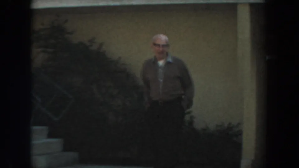 In 1961 Los Angeles home-movie footage, a man in glasses stands near shrubs and exterior stairs beside a sunlit building.