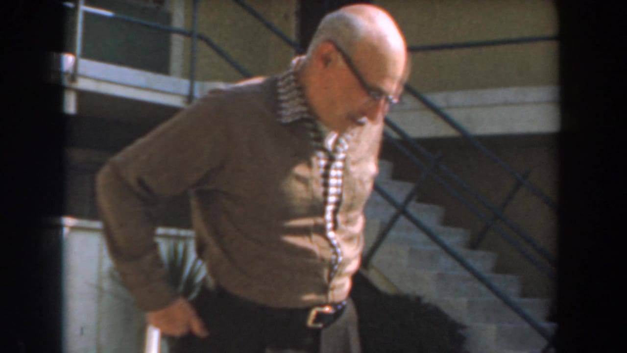 Older man in a cardigan stands beside stairs, looking down toward the ground.