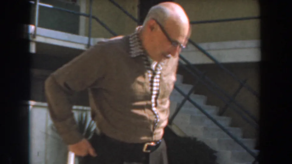 Older man in a brown cardigan looks downward near stairs in 1961 Los Angeles 8mm home-movie footage.