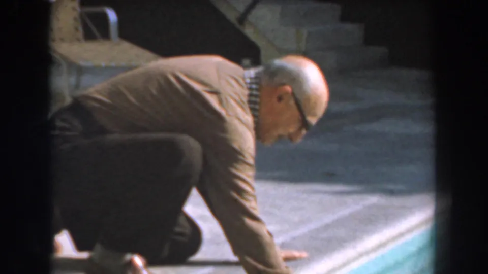 Older man kneels on the concrete pool deck with one hand near the edge in 1961 Los Angeles footage.