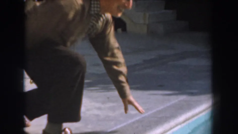 Older man crouches at the pool edge and reaches downward in 1961 Los Angeles 8mm home-movie footage.