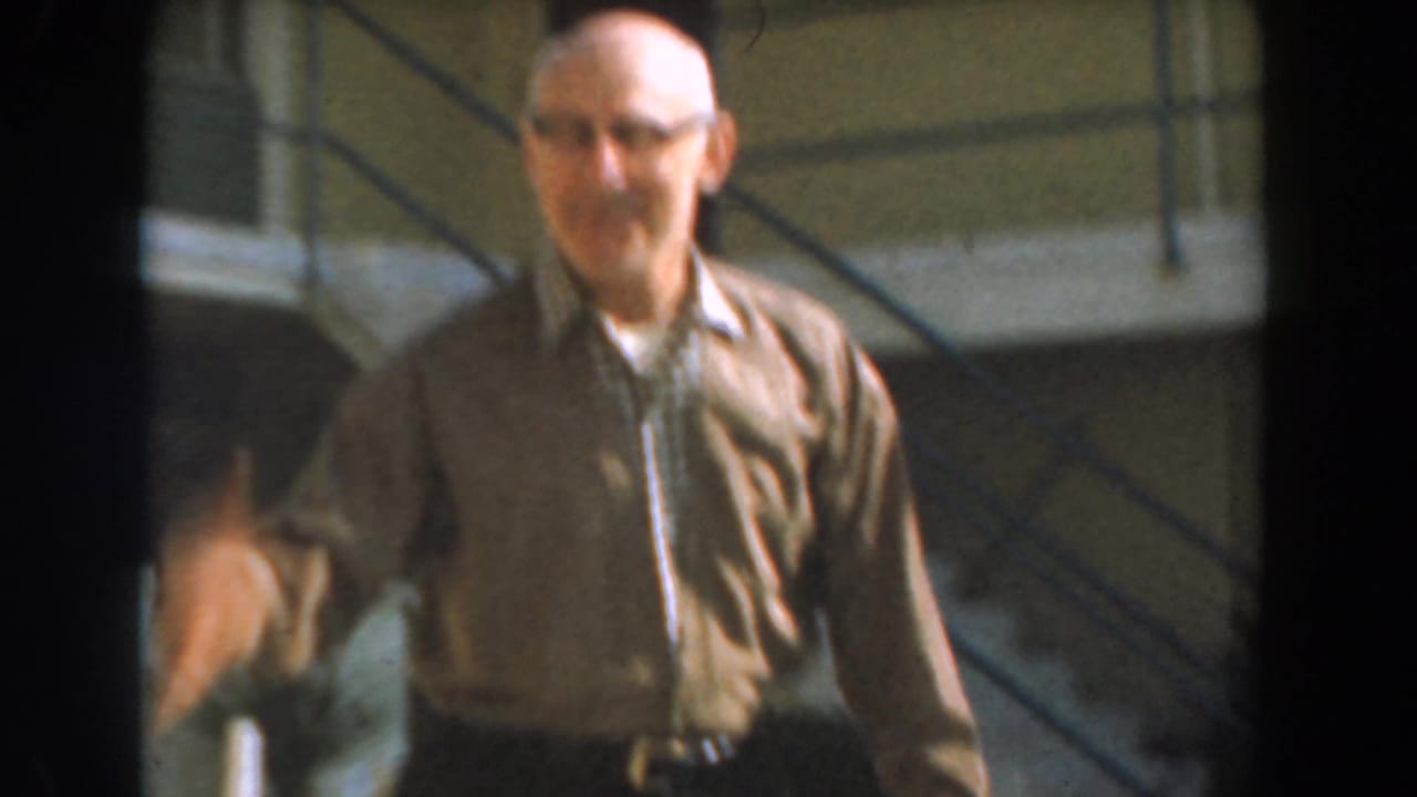 Older man in glasses walking outdoors near a residential building and stairs.
