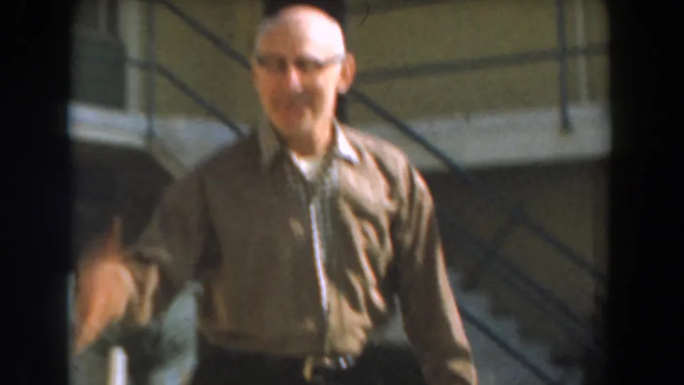 Older man in glasses and a brown jacket appears outside in 1961 Los Angeles home-movie footage, with stairs and building details behind him.