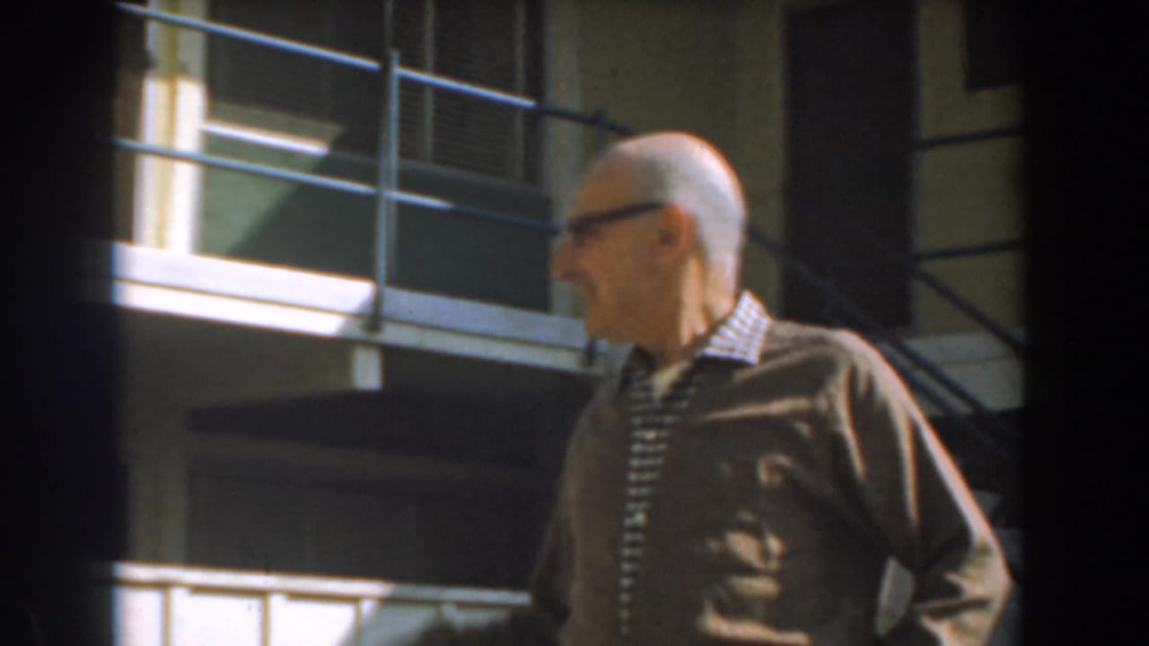 Older man in glasses turns sideways in front of a building balcony and railings.