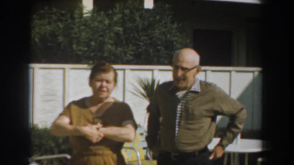 Older couple stands in a Los Angeles backyard, with the woman crossing her arms and the man beside her near a white fence and patio furniture.
