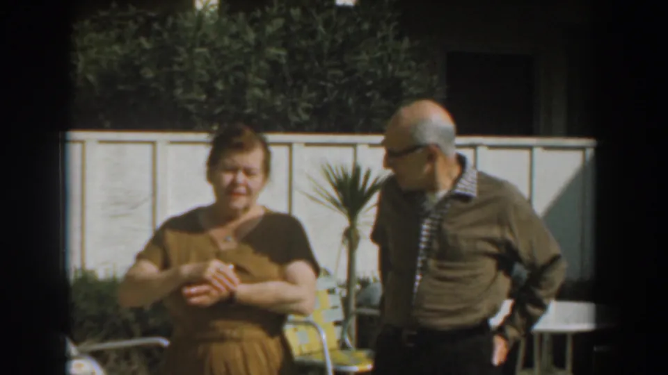 Older couple remains in the sunlit Los Angeles backyard as the man looks toward the woman, with fence, plants, and patio furniture visible.