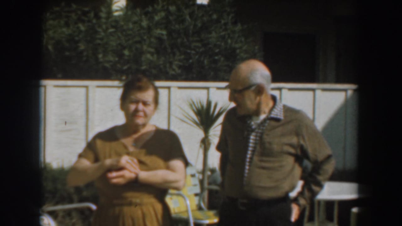Older woman and man standing in a sunny backyard near a white fence.