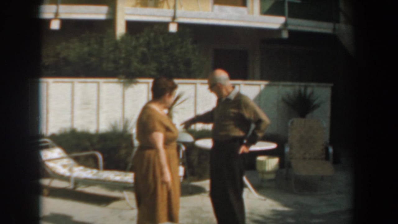 Elderly woman and man stand near patio furniture in a sunny backyard.