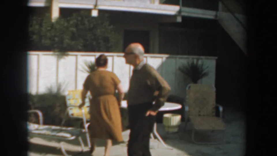 The couple stands near mid-century outdoor chairs and a light fence, both partly turned away in the backyard scene.