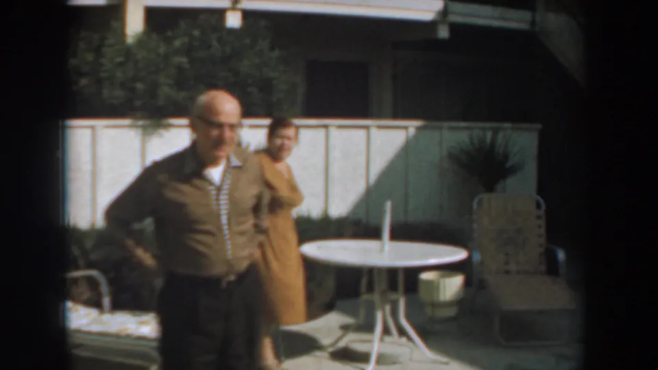 The elderly man moves closer to camera while the woman remains behind him near the round table and lounge chair.