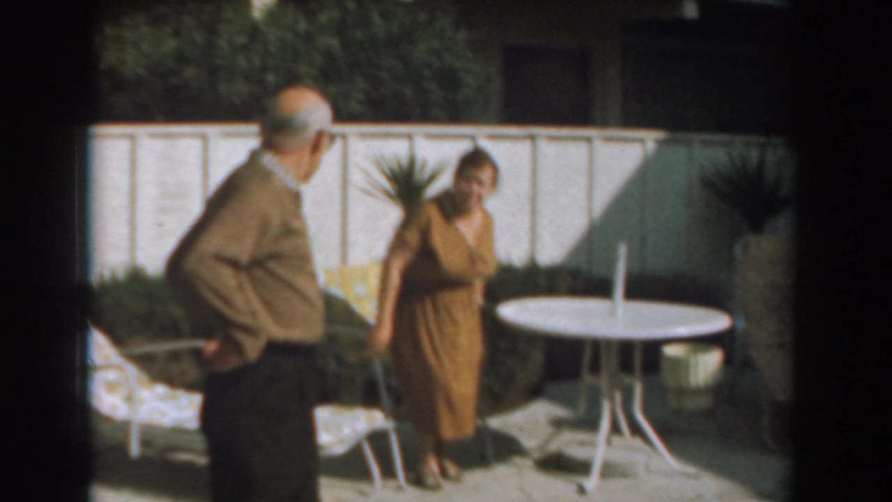 Older man stands near a woman by a patio table in a sunny backyard.