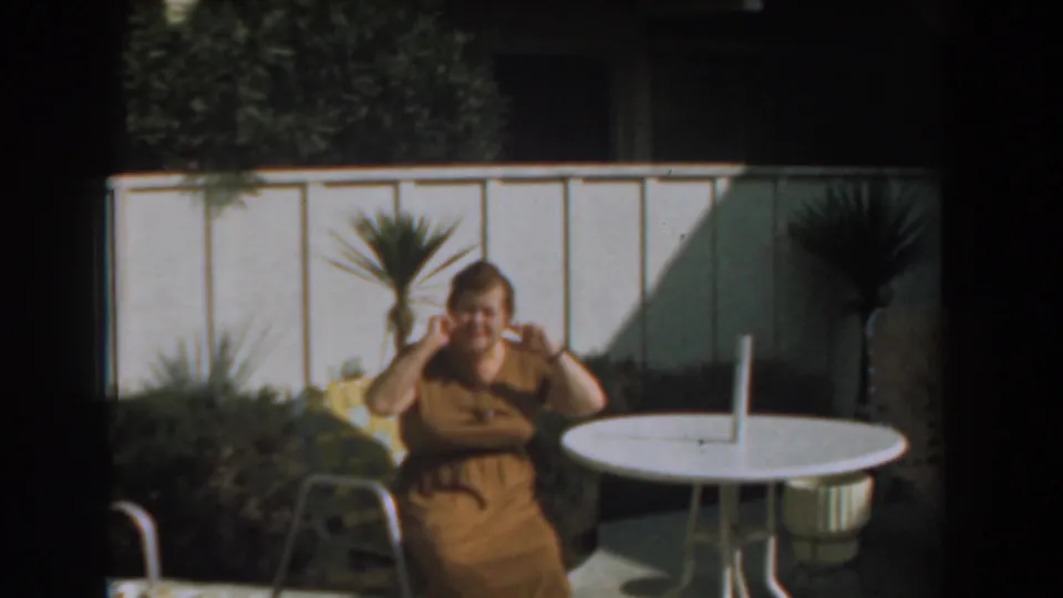 1961 Los Angeles 8mm frame showing a woman smiling and raising her hands while seated beside a backyard patio table.