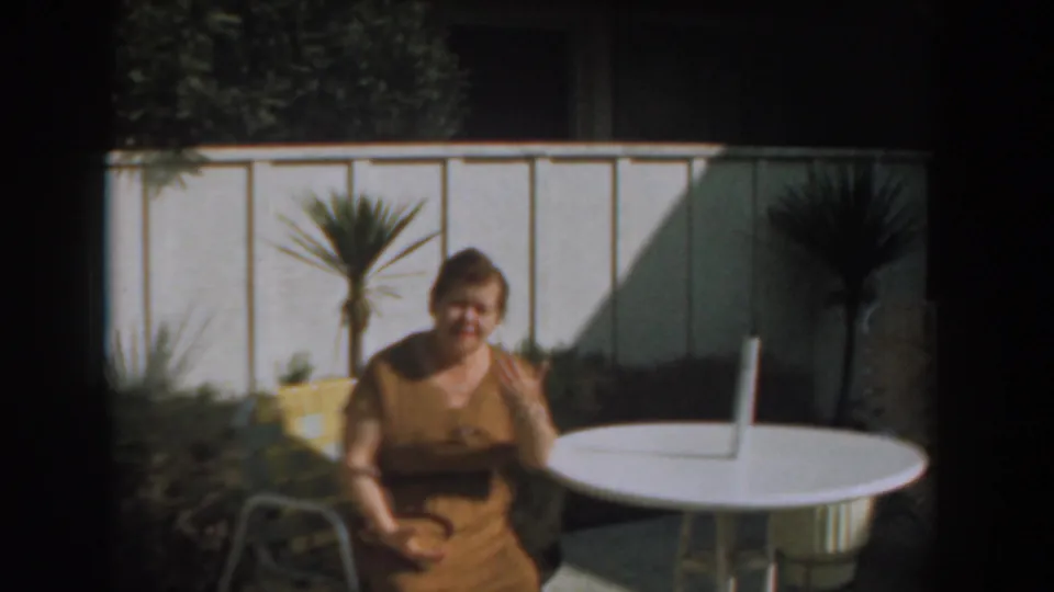 1961 Los Angeles home-movie frame showing a woman waving from a patio chair beside a white round table.