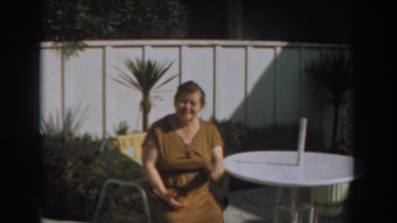 In 1961 Los Angeles home-movie footage, a woman in a brown dress sits near a white patio table, fence, yellow chair, and potted palm.