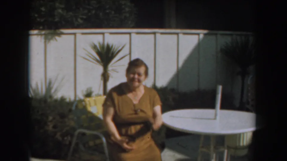 In 1961 Los Angeles home-movie footage, a woman in a brown dress sits near a white patio table, fence, yellow chair, and potted palm.