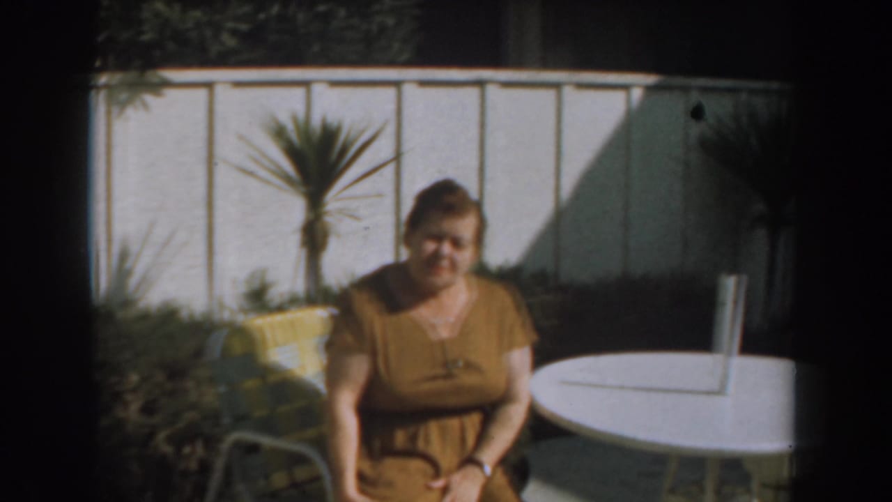 Woman seated outdoors near a white table with a fence and potted palm behind her.