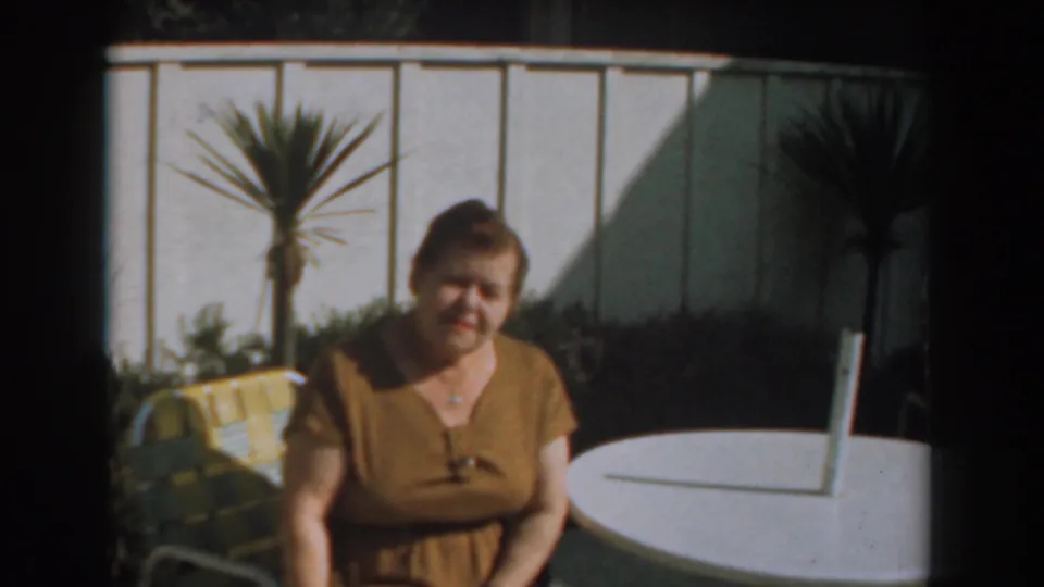 A handheld 8mm frame shows the woman seated on the patio, leaning slightly as sunlight falls across the table, fence, and plants.