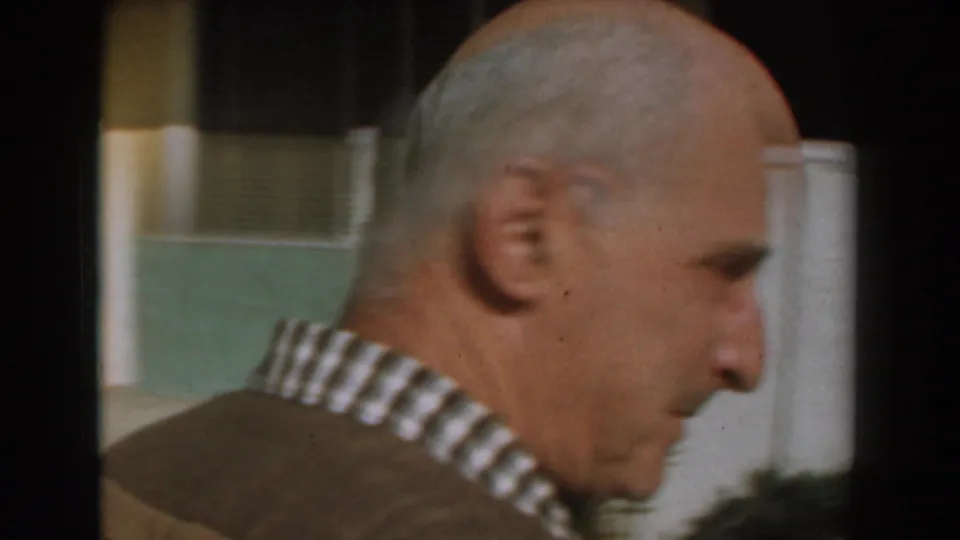 An older man in a brown sweater and plaid shirt passes close to the camera in profile, with the backyard fence and plants behind him.