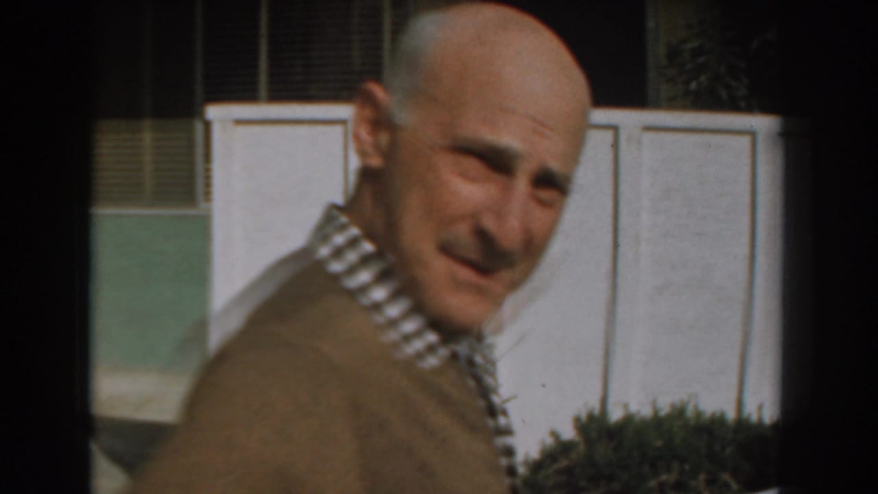 Older bald man in a brown sweater turns toward the camera outdoors near a white paneled wall.
