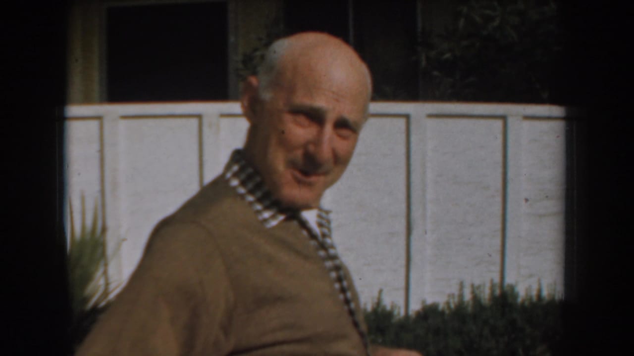 The older man in a brown sweater appears mid-step outdoors, smiling faintly toward the camera against a white wall and planting.