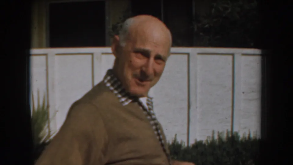 The older man in a brown sweater appears mid-step outdoors, smiling faintly toward the camera against a white wall and planting.