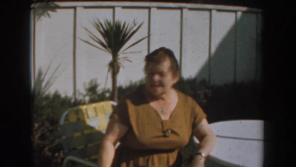 An older woman in a brown dress is seen outdoors near yellow patio seating, white wall panels, shrubs, and a small palm plant.