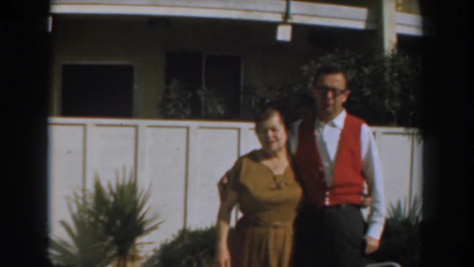 A woman in a brown dress stands close to a younger man in glasses, white shirt, tie, and red vest outside a paneled building.