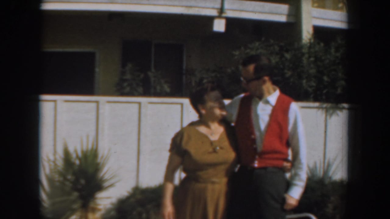 In 1961 Los Angeles home-movie footage, a woman and man stand close together in front of a white fence and apartment building.