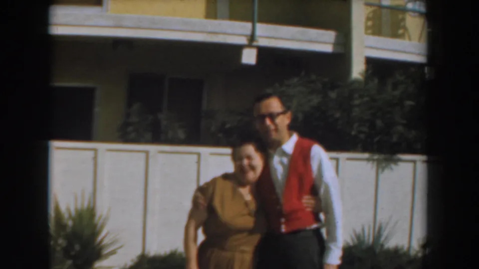 The woman smiles while holding the man's arm in a 1961 Los Angeles apartment exterior scene.