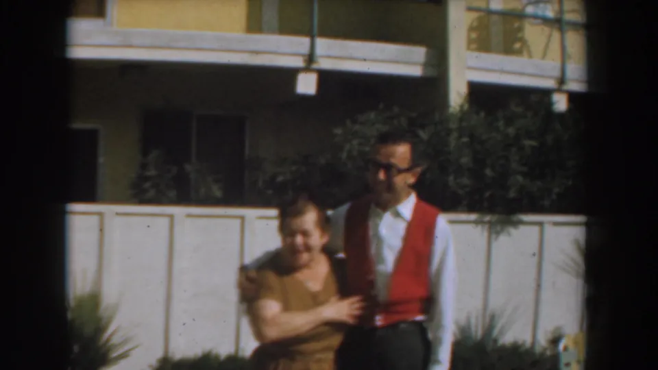 The woman holds the arm of a man in a red vest as they stand outside a Los Angeles residence in 1961 footage.