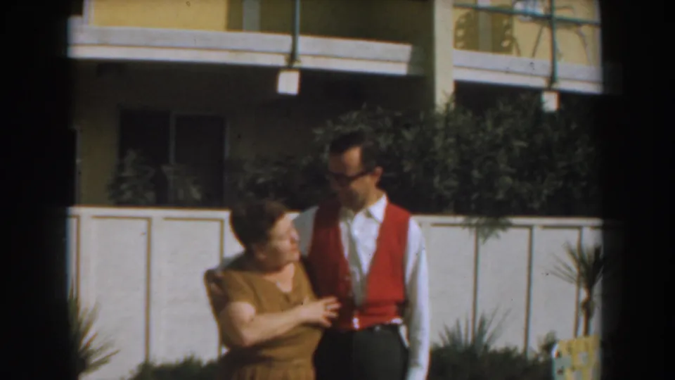 A man and woman interact casually outside a residential building, with the woman gesturing near his red vest.