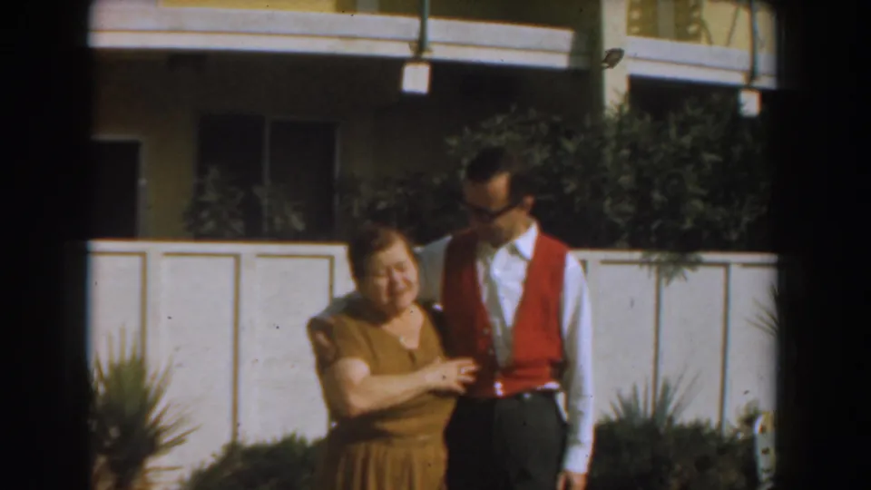 The woman smiles beside the man in a red vest, framed by fencing, shrubs, and a residential exterior in 1961 Los Angeles.