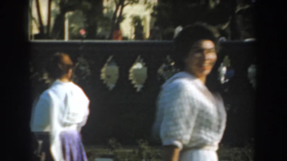 A smiling woman in a white cardigan passes close to the camera, with another adult partly visible near the garden wall.