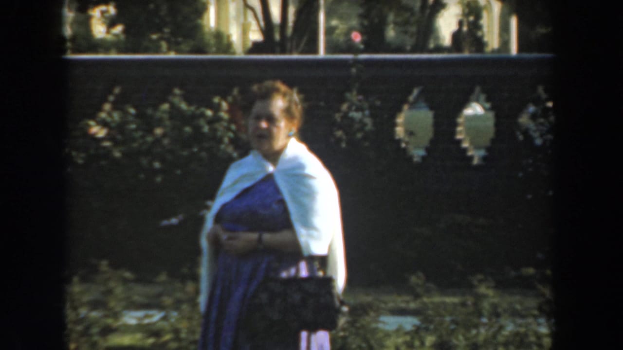Woman in purple dress and white sweater stands near a decorative stone fence.