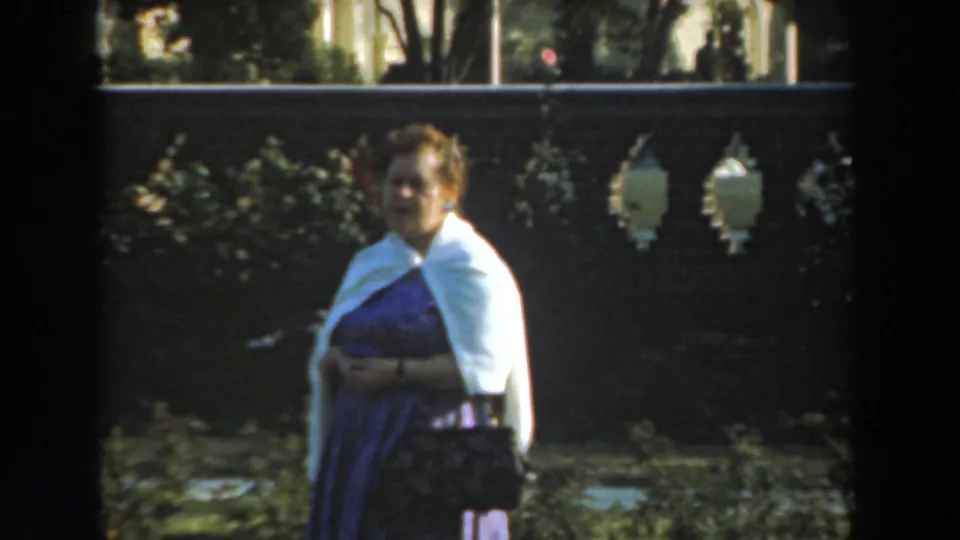 In 1961 Los Angeles home-movie footage, a woman in a purple dress and white sweater stands in a sunlit garden near a gray stone fence.