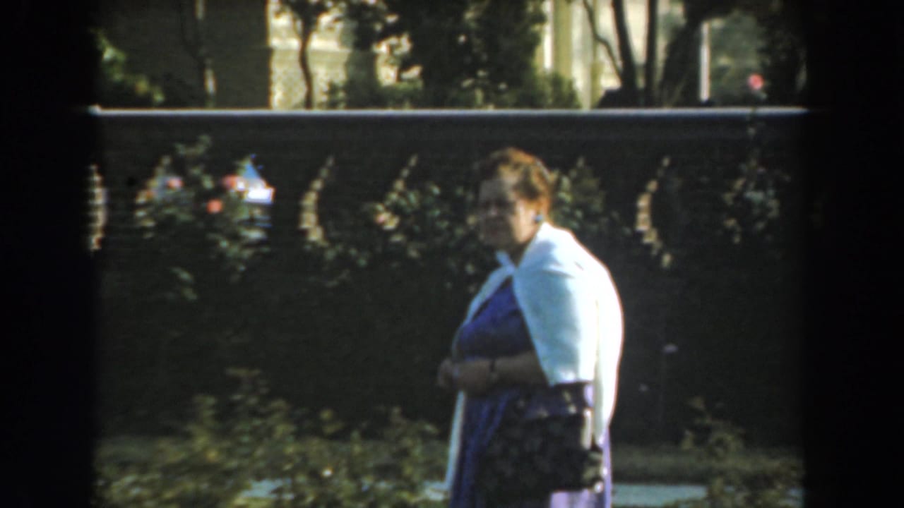 Woman holds a dark handbag while standing beside foliage and a stone fence.