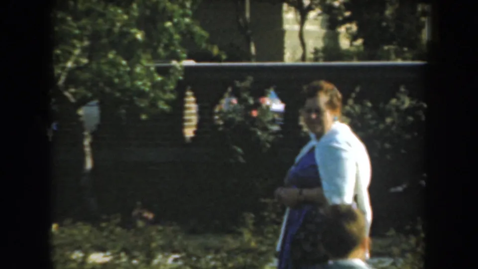 The woman turns slightly in the Los Angeles garden scene, with foliage and the decorative fence behind her and a partial head at the lower frame edge.