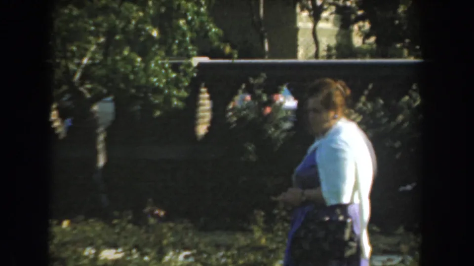 The woman is seen more in profile, standing beside garden foliage and the decorative gray fence in the 1961 Los Angeles home-movie clip.