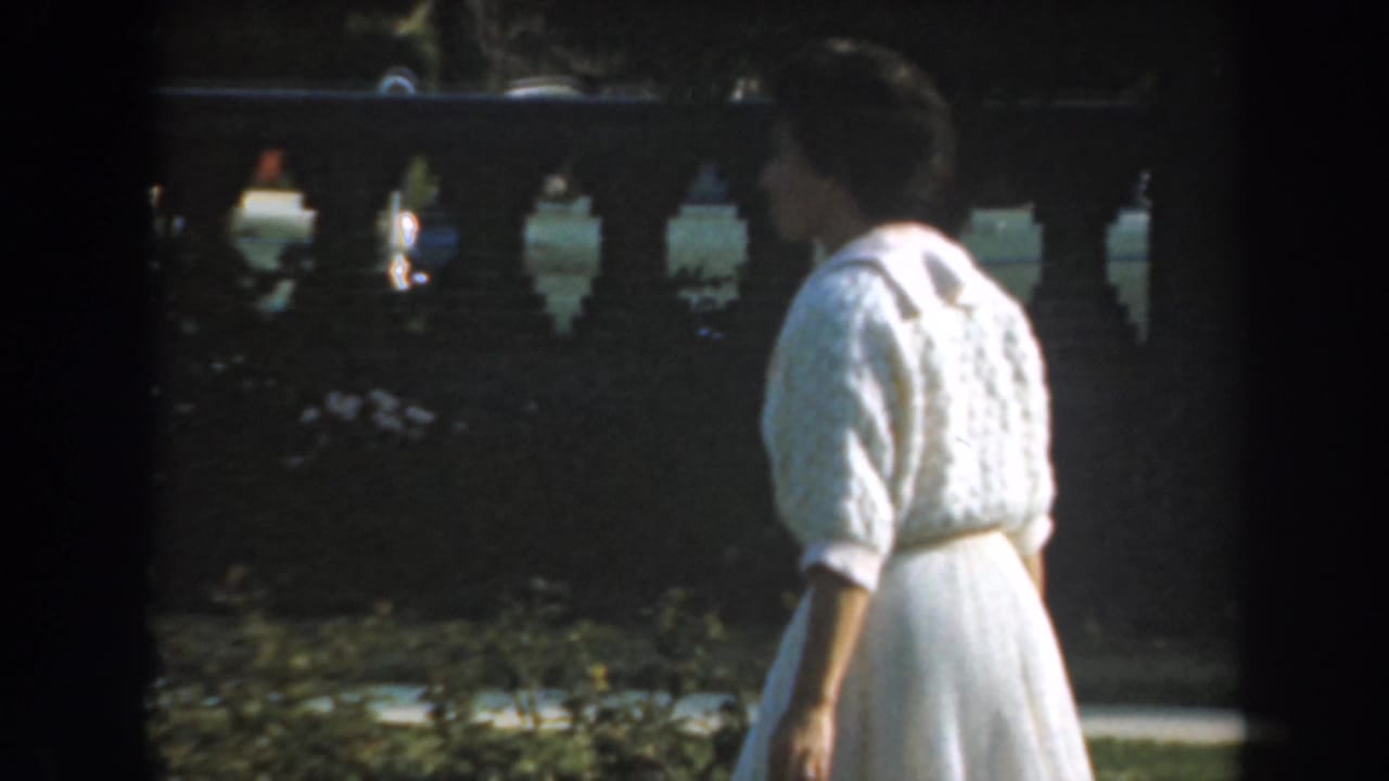 Woman in a white dress and cardigan walks beside a garden balustrade.