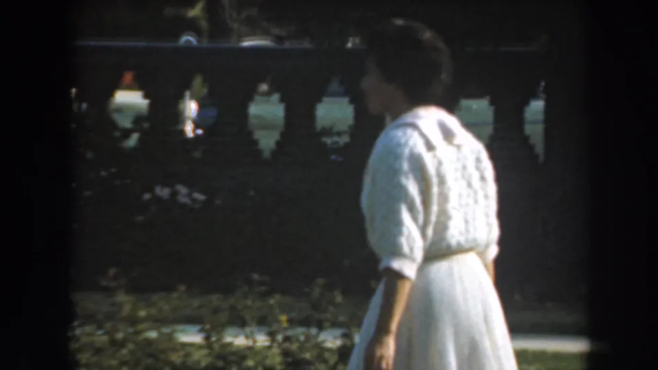 A woman in a white dress and knitted cardigan walks through a Los Angeles garden, seen in profile against a balustrade.