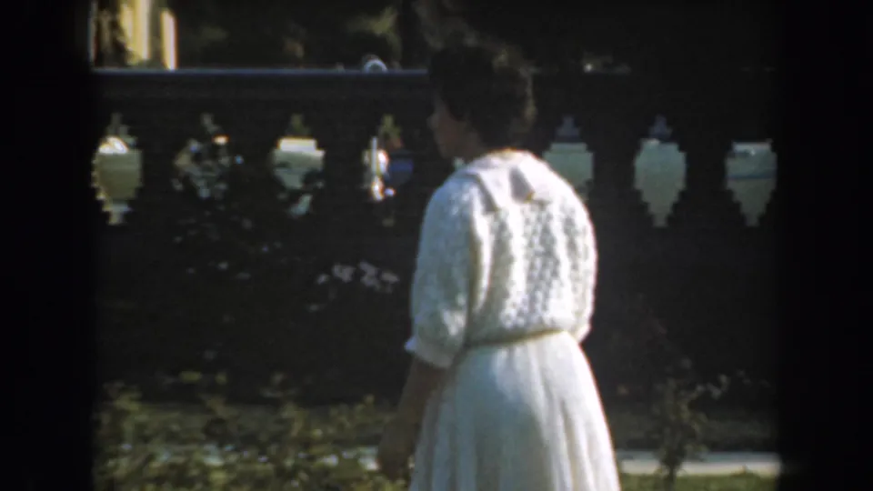 From behind, the woman in a white dress and cardigan walks near a decorative balustrade in the 1961 Los Angeles garden footage.