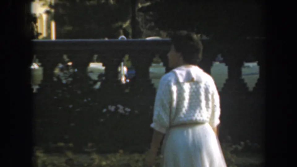 The woman in a white dress appears near shrubs and a decorative balustrade in soft, sunlit 8mm footage from Los Angeles.