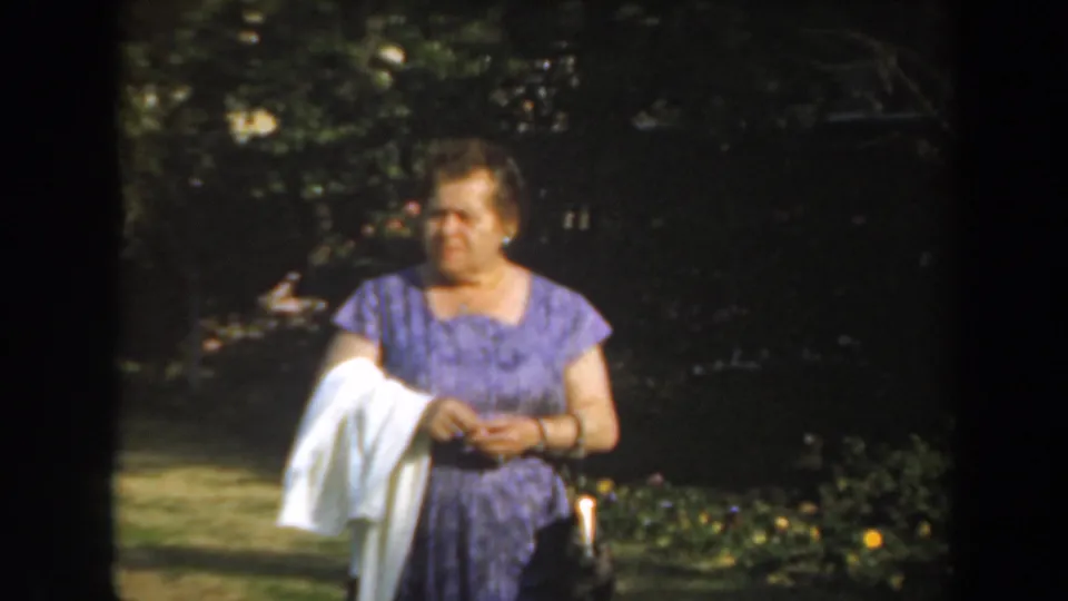 In 1961 Los Angeles home-movie footage, a woman in a purple patterned dress holds a white cloth in a sun-dappled garden.
