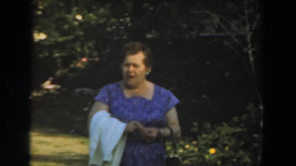 A woman in a purple patterned dress moves through a Los Angeles garden, carrying a white cloth over her arm.